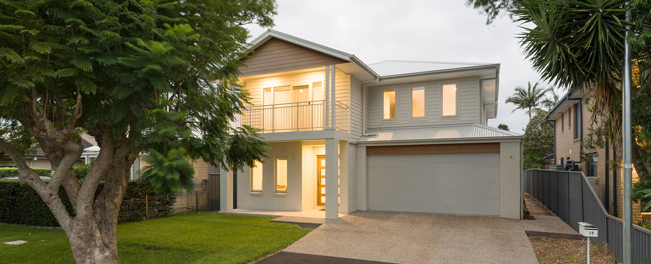 Modern two-story residential home at dusk with warm interior lighting. The light gray house features a garage, upper-level balcony, and large windows. A mature tree stands in the front yard on the left, with tropical palms visible on the right. A gravel driveway leads to the garage, and neighboring houses are visible on either side.