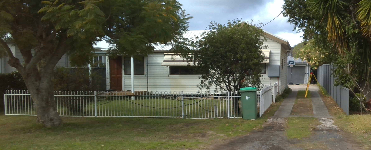 Weathered single-story white house with red door, surrounded by white picket fencing and overgrown vegetation, including large trees and tropical plants. A green waste bin sits near the fence, with a gravel driveway leading to the property and neighboring structures visible on either side.