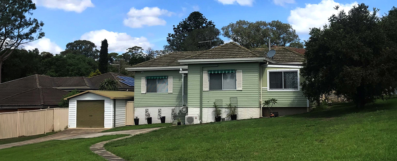 Single-story residential house in Campbelltown with green and white weatherboard exterior, tiled roof, front garage, manicured lawn, brick pathway, and mature trees in background under blue sky with white clouds.