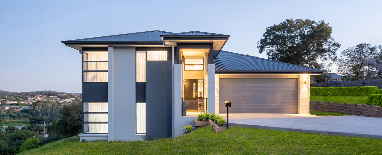 Modern two-story house with dark blue and white exterior, metal roof, large windows, and attached garage, situated on landscaped green lawn at dusk with warm interior lighting and suburban hillside landscape visible in background.