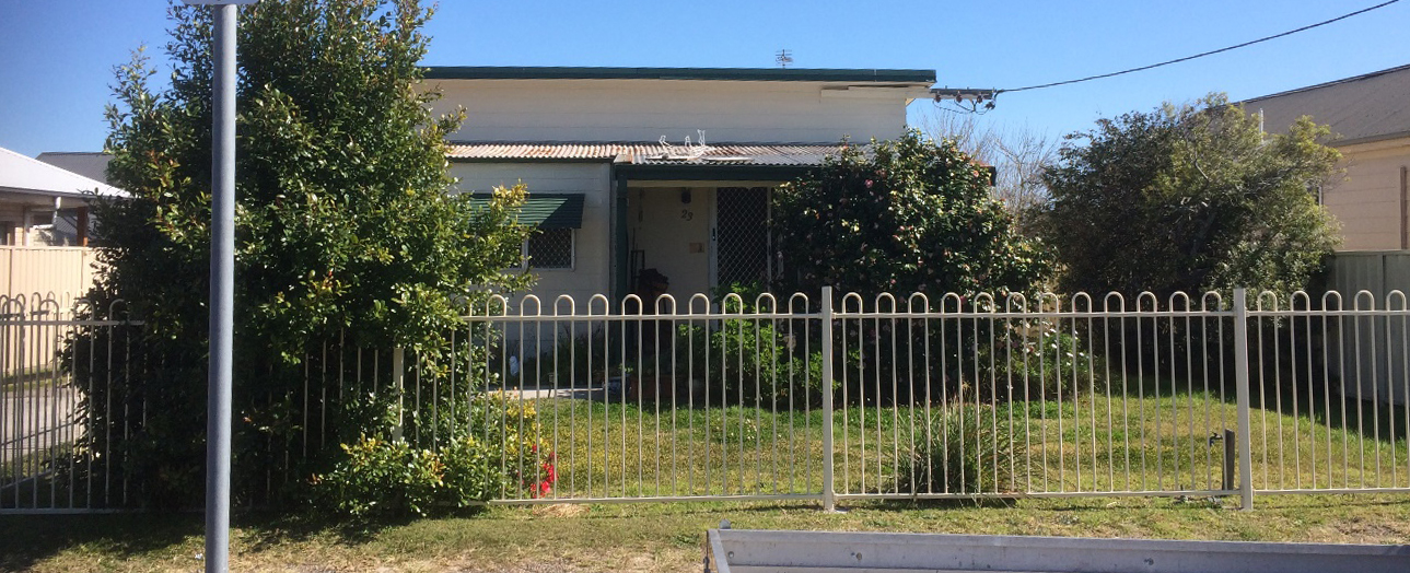 A single-story residential house with weathered cream and green exterior, surrounded by overgrown vegetation and enclosed by a white metal fence. The front yard is unkempt with dry grass and wild plants growing around the property. Clear blue sky visible above.