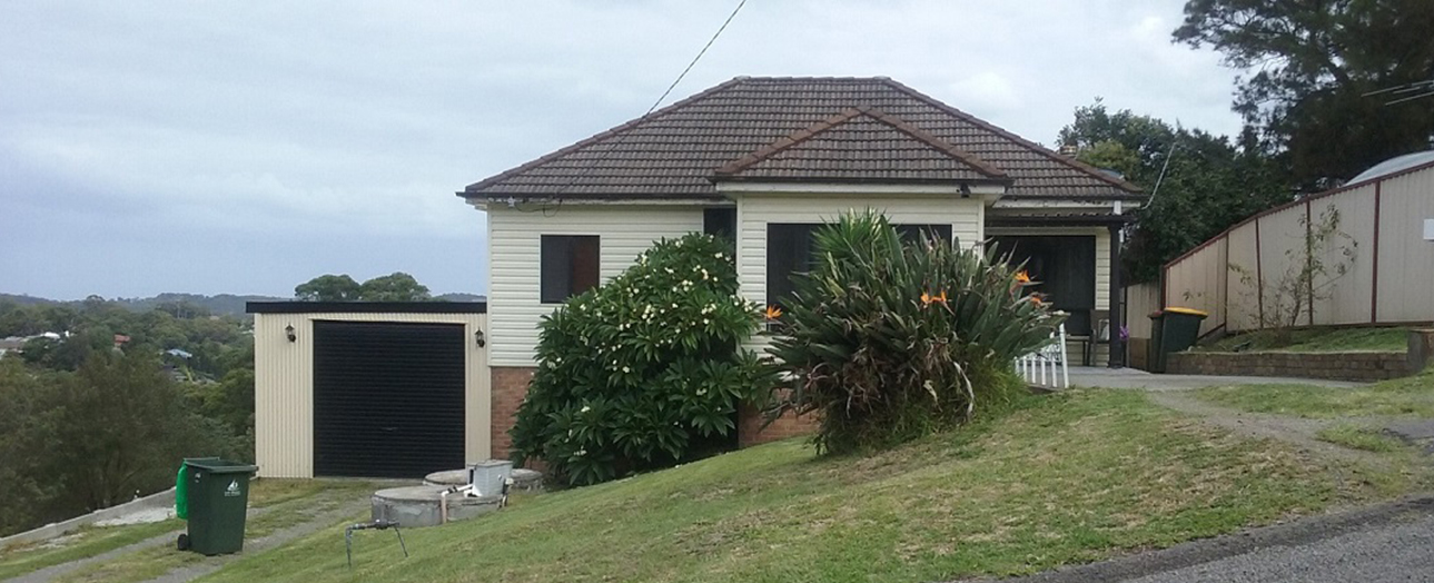 Single-story cream-colored house with dark tile roof and brick base, featuring a detached garage on the left. The front yard has overgrown grass and tropical plants including a large palm tree. Green waste bin visible in foreground. Rural hillside setting with trees and fencing in background.