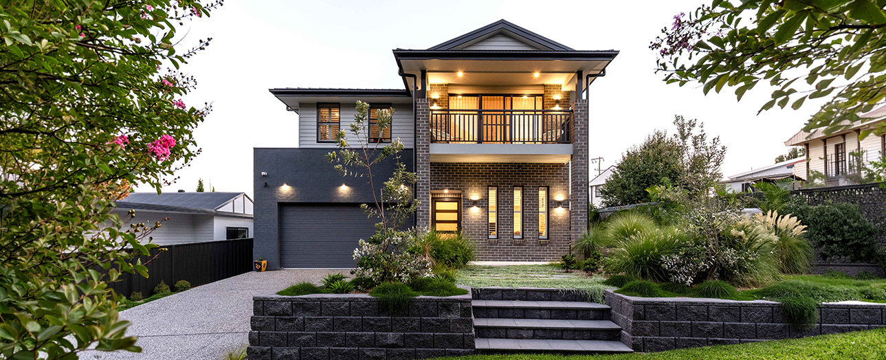 Modern two-story brick house in Charlestown with dark gray facade, illuminated windows, and wooden upper-level balcony. Front features a dark garage door, landscaped garden with ornamental grasses and flowering shrubs, black stone retaining walls, gravel driveway, and manicured lawn. Evening lighting highlights the contemporary architectural design.
