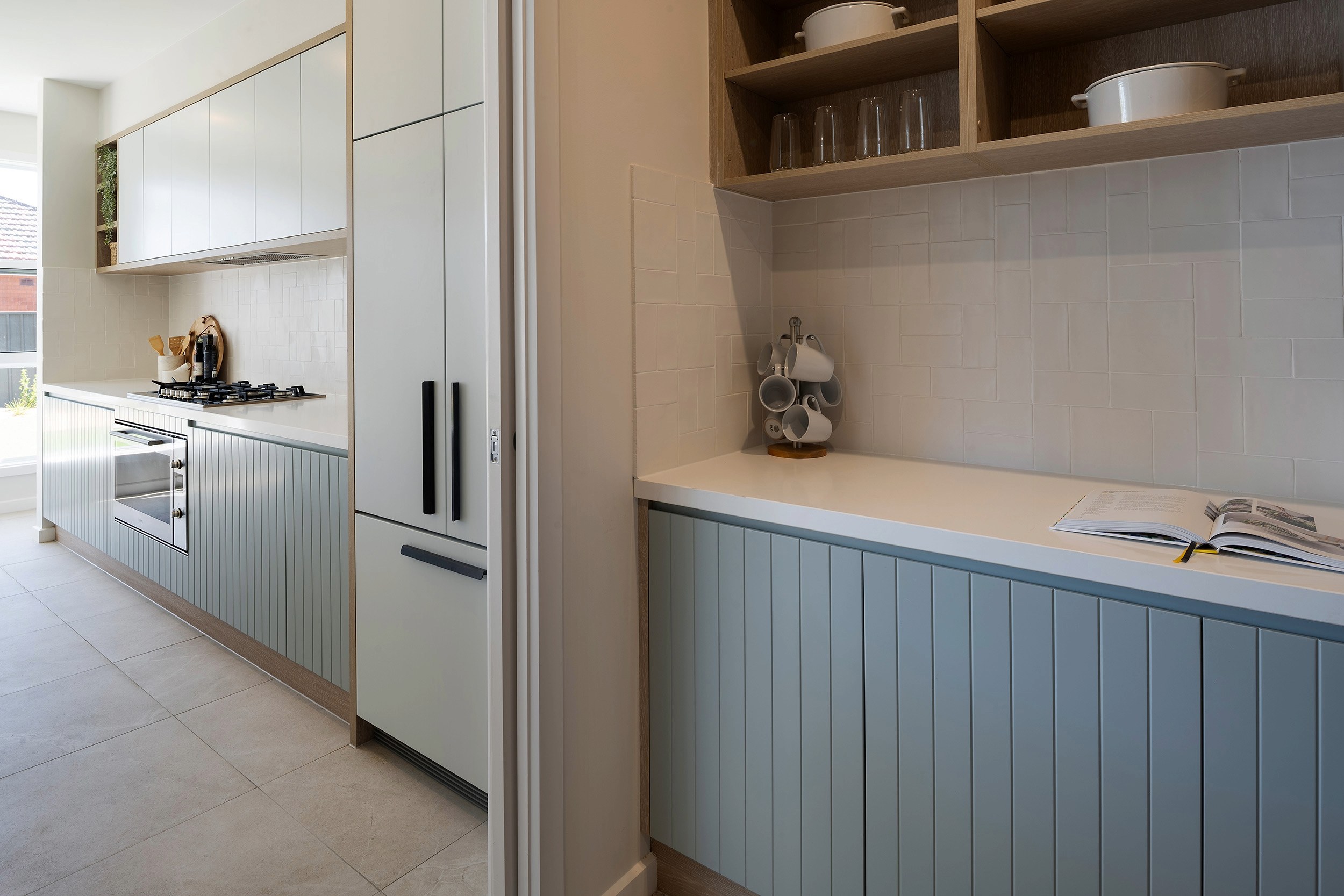 Modern coastal kitchen with white upper cabinets and blue-green vertical paneled lower cabinets. Left side features a gas cooktop, built-in oven, and stainless steel refrigerator. Right side shows a prep counter with white subway tile backsplash, open wooden shelving with dishware, and a mug tree. Natural light streams through a window, and the space has light tile flooring.