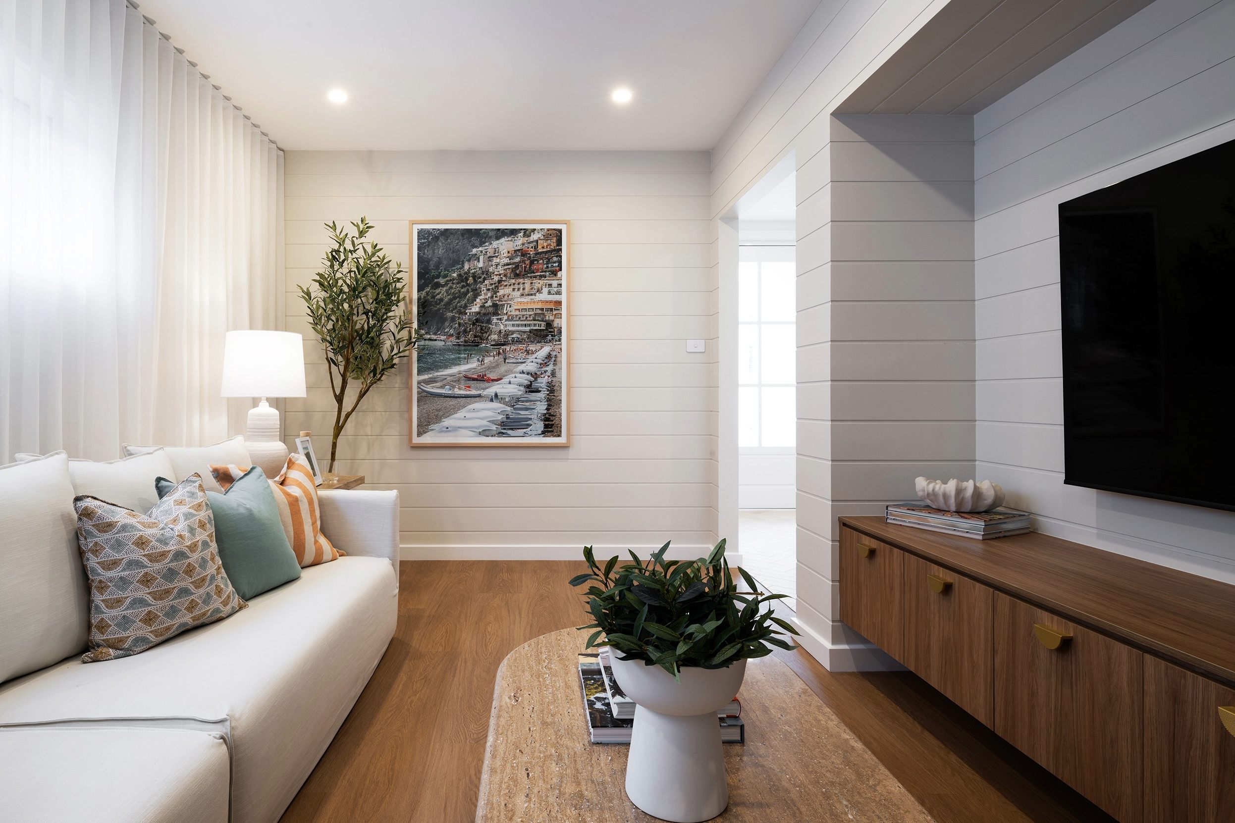 Modern living room with white shiplap walls, cream sofa with patterned pillows, wooden flooring, and a framed coastal photograph depicting a cliffside Italian village. A potted plant sits on a round table in the foreground, with a wall-mounted TV and wooden media console on the right, and floor-to-ceiling curtains on the left.