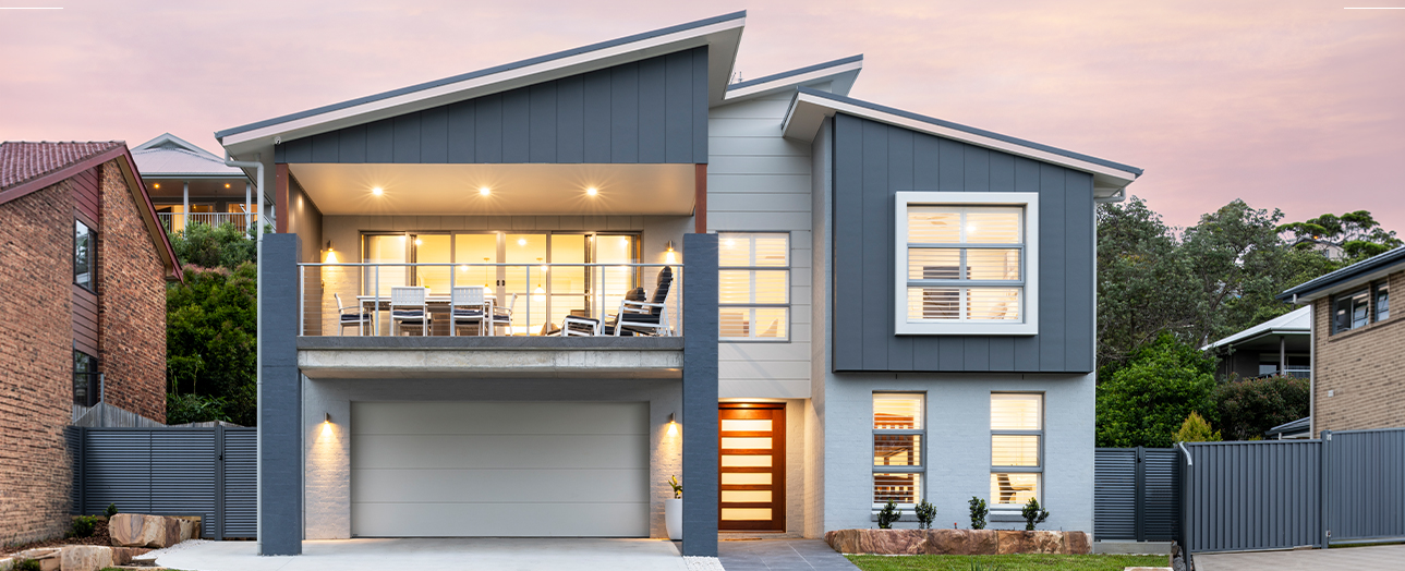 Modern two-story house with blue and white exterior, featuring a double garage, illuminated balcony on upper level, large windows, and horizontal wood slat front door. Dusk sky with pink tones in background. Neighboring brick houses visible on either side.