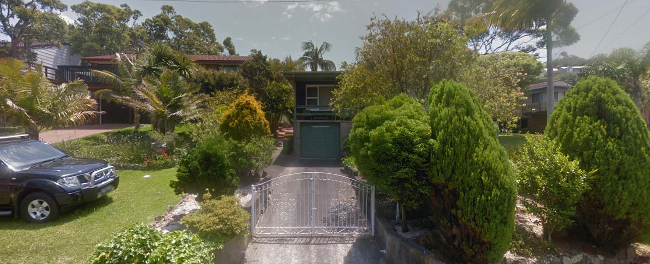 Suburban residential property with overgrown landscaping. A dark-colored SUV is parked on the left lawn. A metal driveway gate leads to a two-story house with dark green garage doors, partially obscured by dense green hedges and trees. The property features palm trees, mature vegetation, and manicured lawn. Power lines are visible overhead.