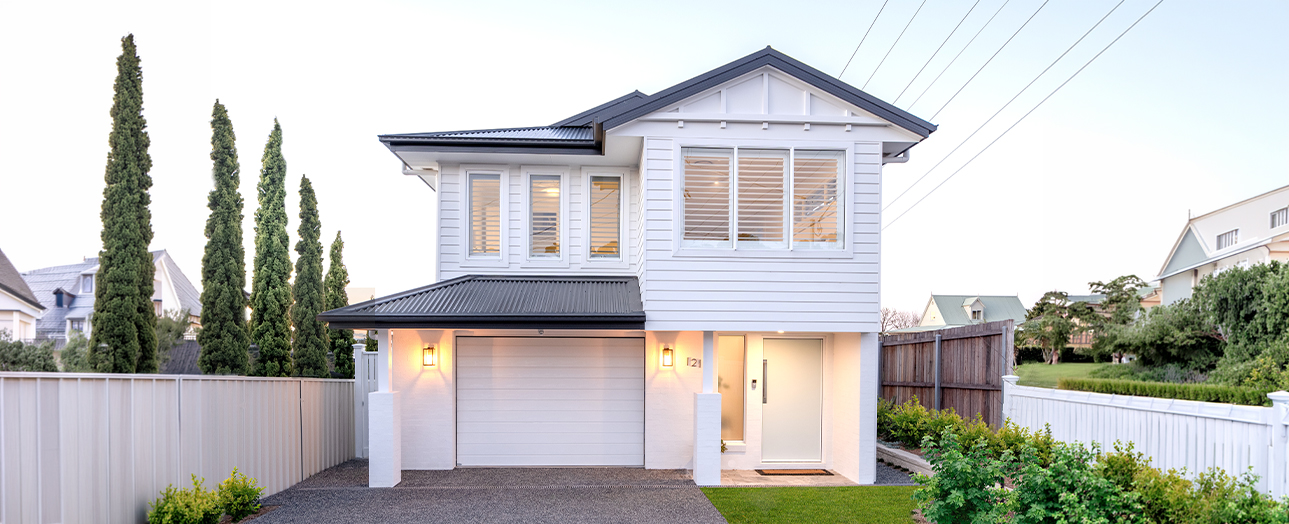 Modern two-story residential house with white horizontal siding and dark roof, featuring a white garage door, illuminated entry, large upper-level windows, tall cypress trees on left side, white fence, and manicured landscaping with green lawn.