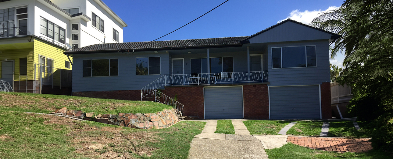 Blue and brick residential house with metal staircase railing leading to upper level, double garage doors below, sloped grassy yard with stone retaining wall, palm tree visible on right, clear blue sky, neighboring yellow house visible on left.