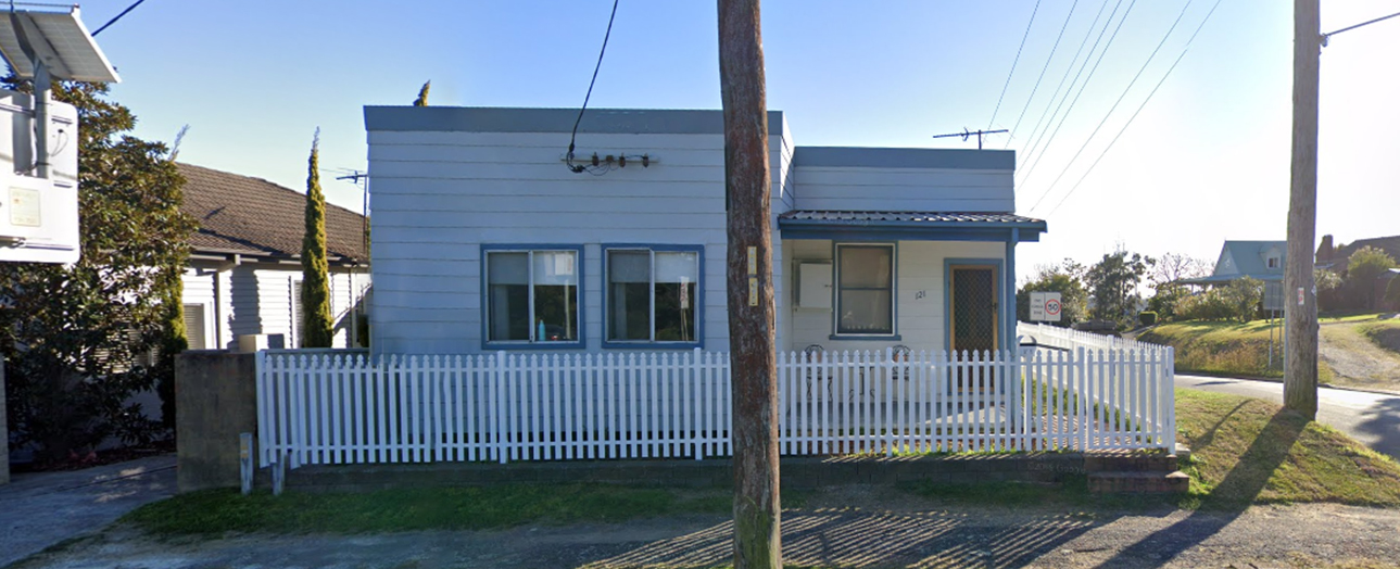 Weathered blue and white cottage with corrugated iron cladding and white picket fence, before renovation. The modest single-story house features two front windows, a small covered porch, and is situated on a residential street with power lines and neighboring homes visible.