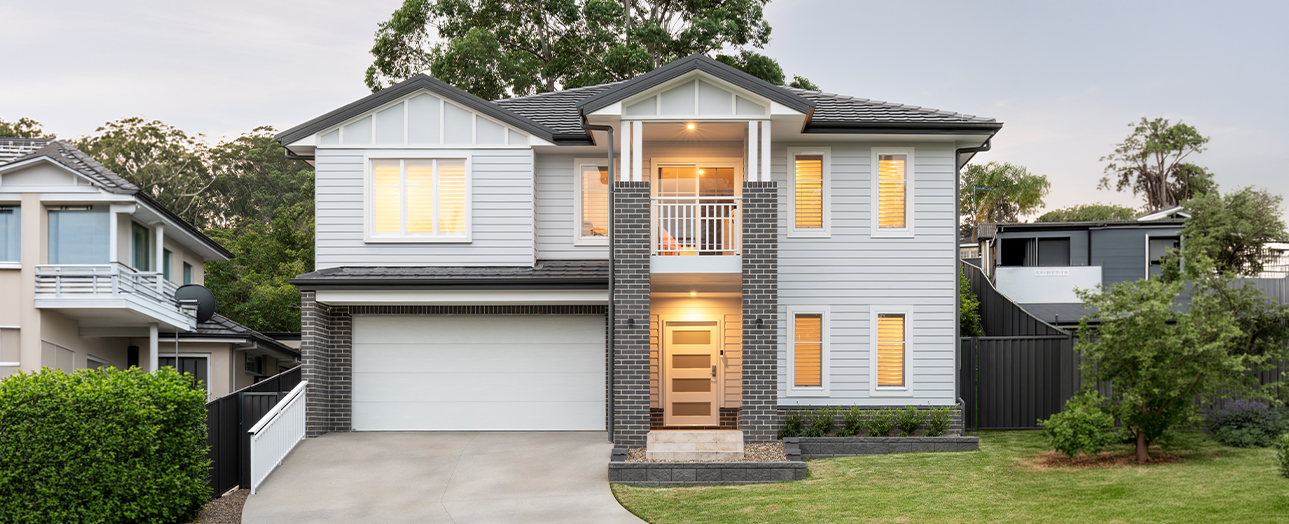 Modern two-story residential house with white and dark brick facade, white garage door, illuminated windows, front porch with railings, manicured lawn, and surrounding trees in a suburban neighborhood setting.