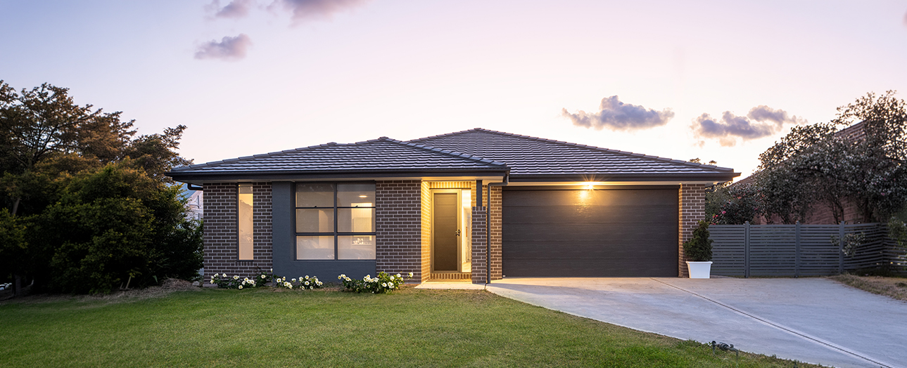 Modern single-story brick house at dusk with illuminated entry and windows, dark hip roof, double garage, concrete driveway, manicured lawn, landscaping with white flowers, and surrounding trees against twilight sky.