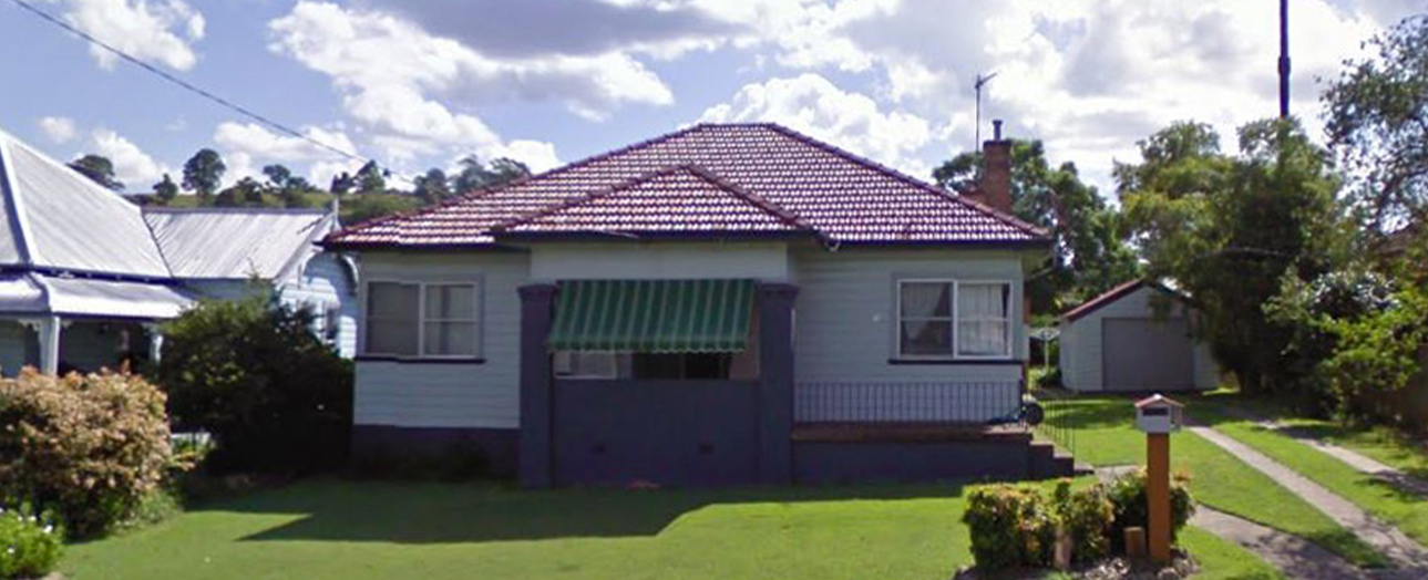 Dungog residential house before renovation, featuring a single-story weatherboard home with red-tiled roof, green awning over central entrance, elevated on stumps with green lawn and surrounding trees under blue sky with white clouds.