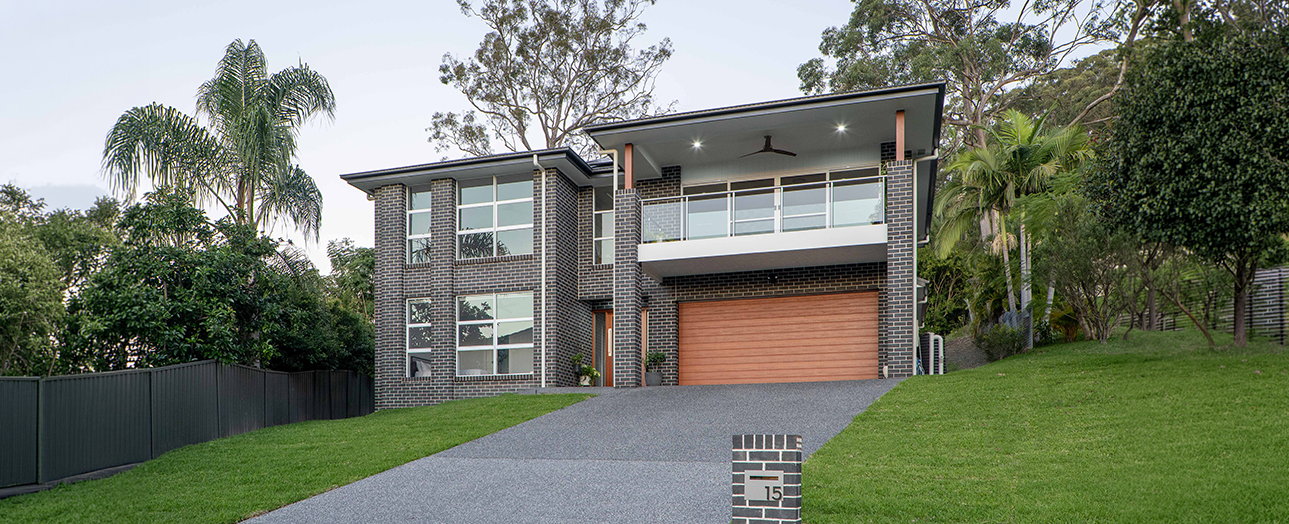 Modern two-story brick house with orange garage door, large windows, and white upper-level balcony, set on sloped green lawn with palm trees and native vegetation, driveway leading to house number 15.