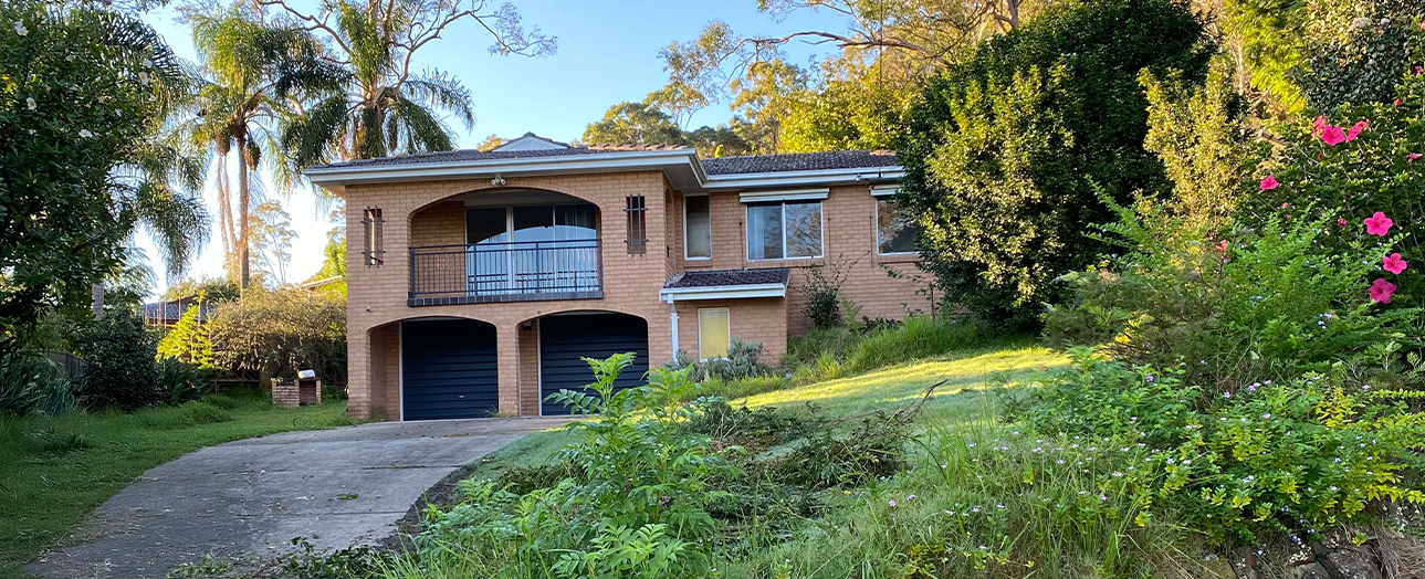 Two-story brick house with overgrown garden and wild vegetation. The property features a curved balcony on the upper level, double garage doors, and is surrounded by dense green shrubs, flowering plants with pink blooms, and mature trees. The landscape appears unmaintained with long grass and tangled plantings covering much of the yard.