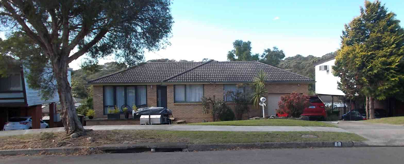 Single-story brick house with dark tiled roof, large front windows, and landscaped garden featuring mature trees and shrubs. Street view showing driveway, parked vehicles, and neighboring properties with hills in background.