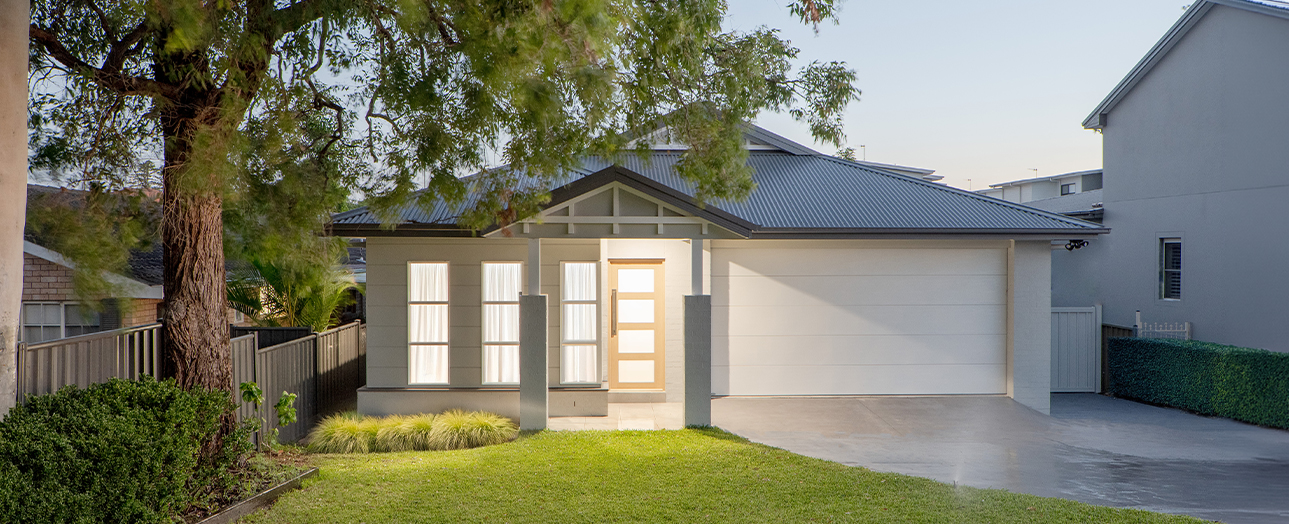 Modern single-story house with cream and gray exterior, metal roof, and large garage door. The front features a gabled entrance with glass panels and wooden door. A manicured lawn, ornamental grasses, and mature tree with drooping branches occupy the front yard. Neighboring houses visible on either side. Clear sky above.