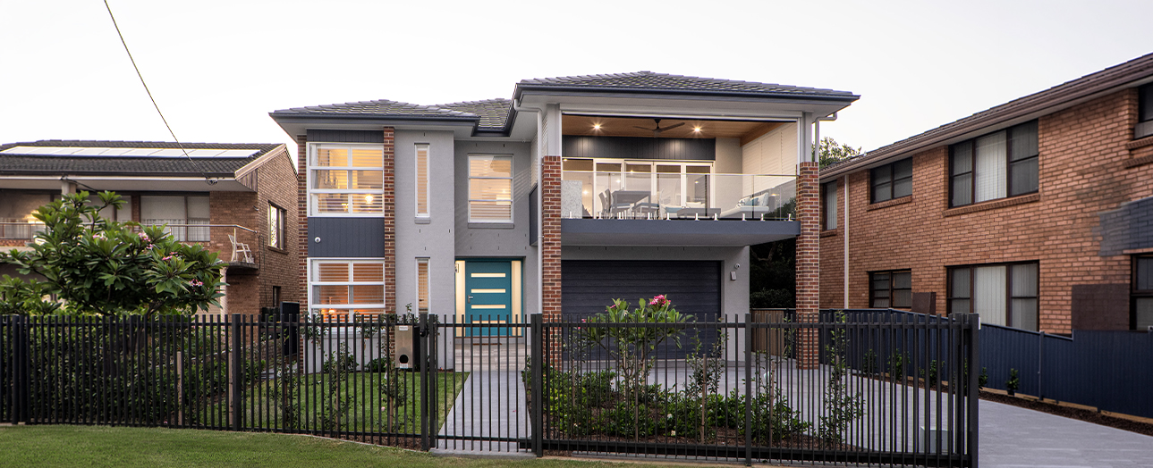 Modern two-story brick residential home with contemporary design features including a teal front door, large windows, dark metal railings, and a garage. The house is surrounded by manicured landscaping with green lawn and flowering plants, enclosed by black metal fencing. Adjacent brick homes visible on either side.