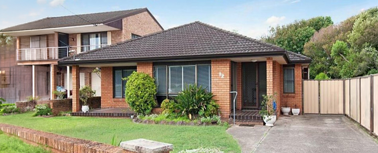 Brick residential home with dark roof and front garden, featuring a single-story section with large windows and a two-story section to the left. The property includes a manicured lawn, potted plants near the entrance, a concrete driveway, and metal fencing along the perimeter, with trees visible in the background under a clear sky.