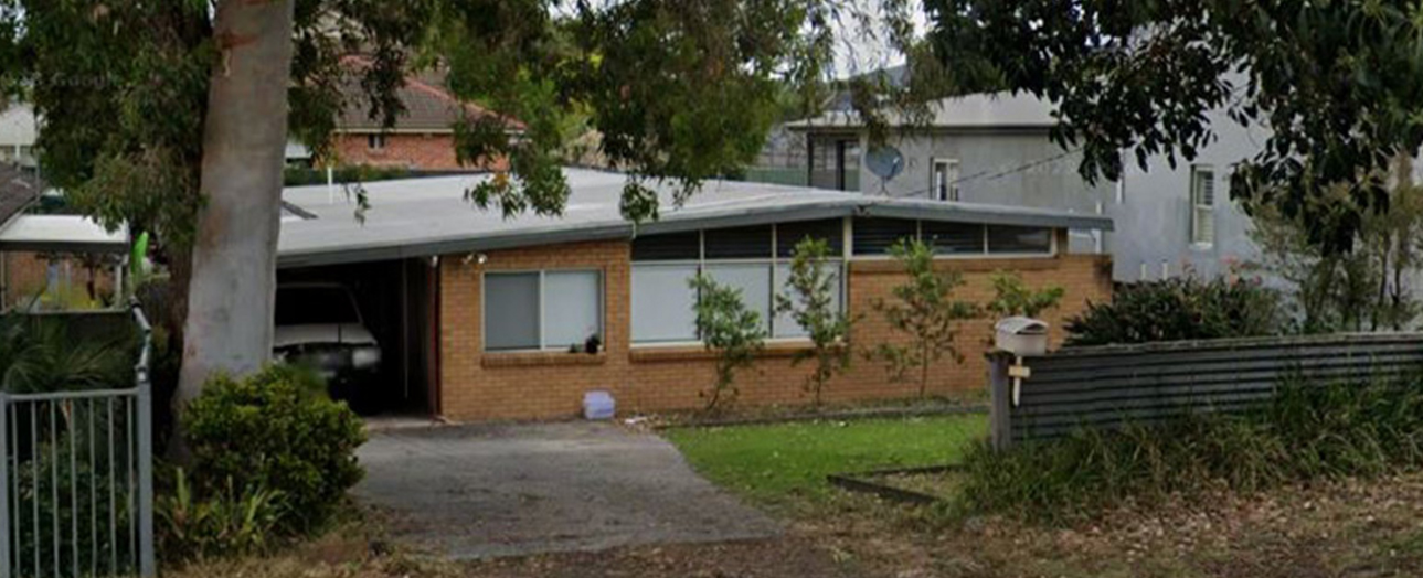 Single-story brick residential home with flat roof, carport, and large windows, surrounded by mature trees and overgrown vegetation, with neighboring houses visible in background.