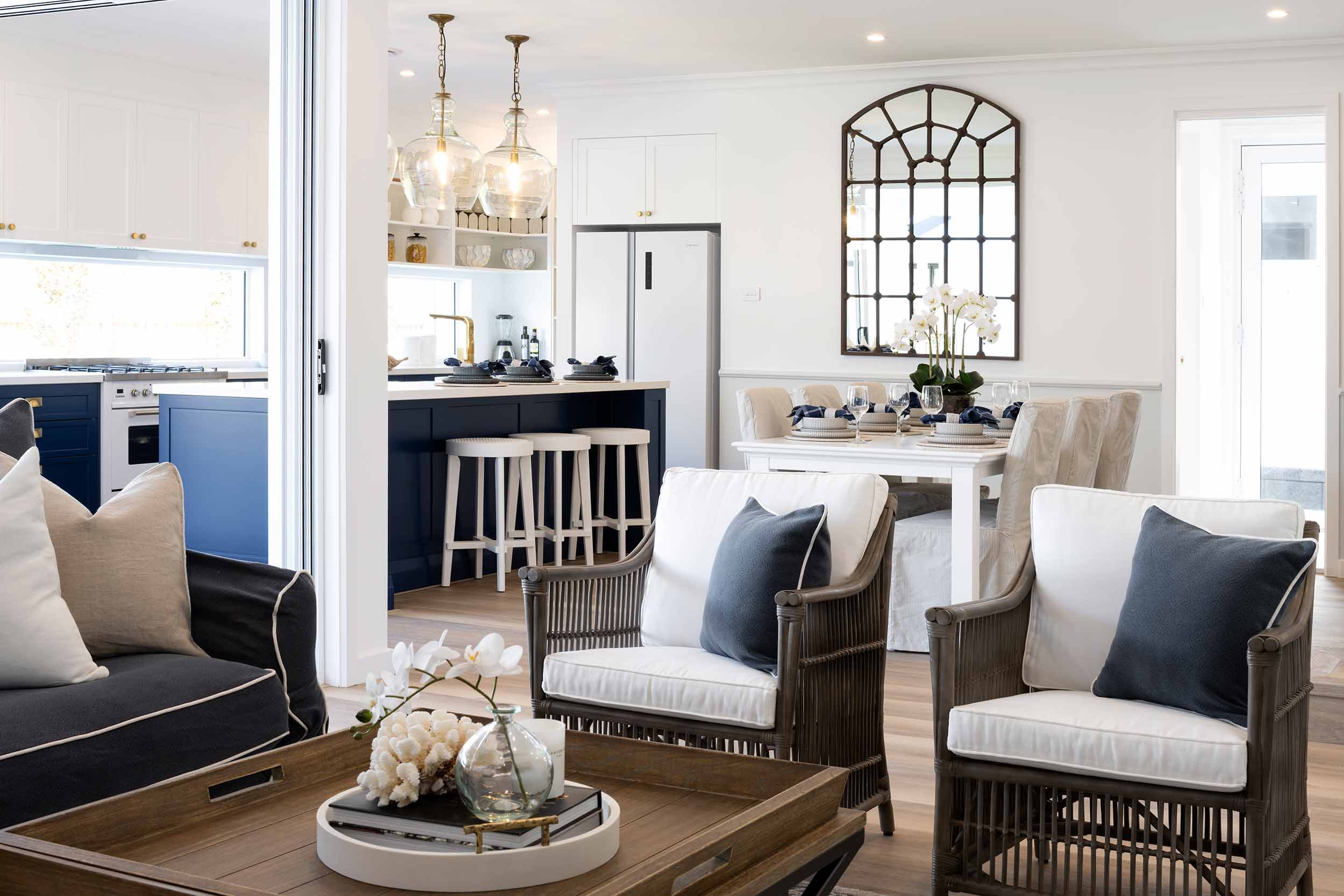 Modern open-plan living space featuring a coastal design with navy blue and white color scheme. Foreground shows wooden armchairs with cream cushions and navy pillows flanking a round coffee table with decorative accessories. Background displays a kitchen with navy cabinetry, white countertops, pendant lighting, and bar seating. A decorative arched mirror hangs above a white console table with white orchids and tableware. Large windows provide natural light throughout the bright, a