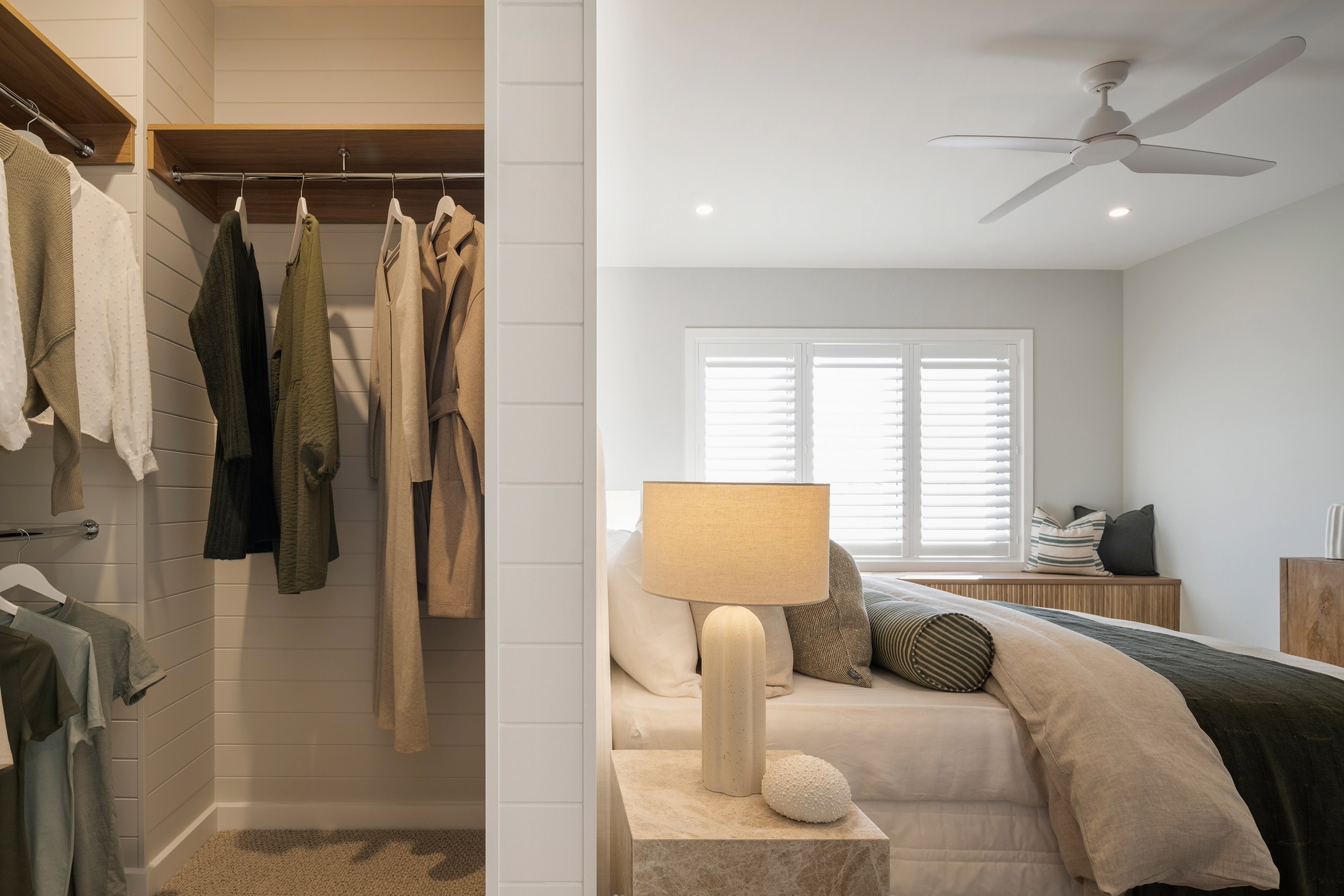 Modern bedroom with open walk-in closet featuring white subway tile walls, hanging neutral-toned clothing, and shelving. Main bedroom area shows bed with cream bedding, decorative pillows, ceramic table lamp, and shuttered windows. Ceiling fan and recessed lighting overhead. Wood dresser visible in background.