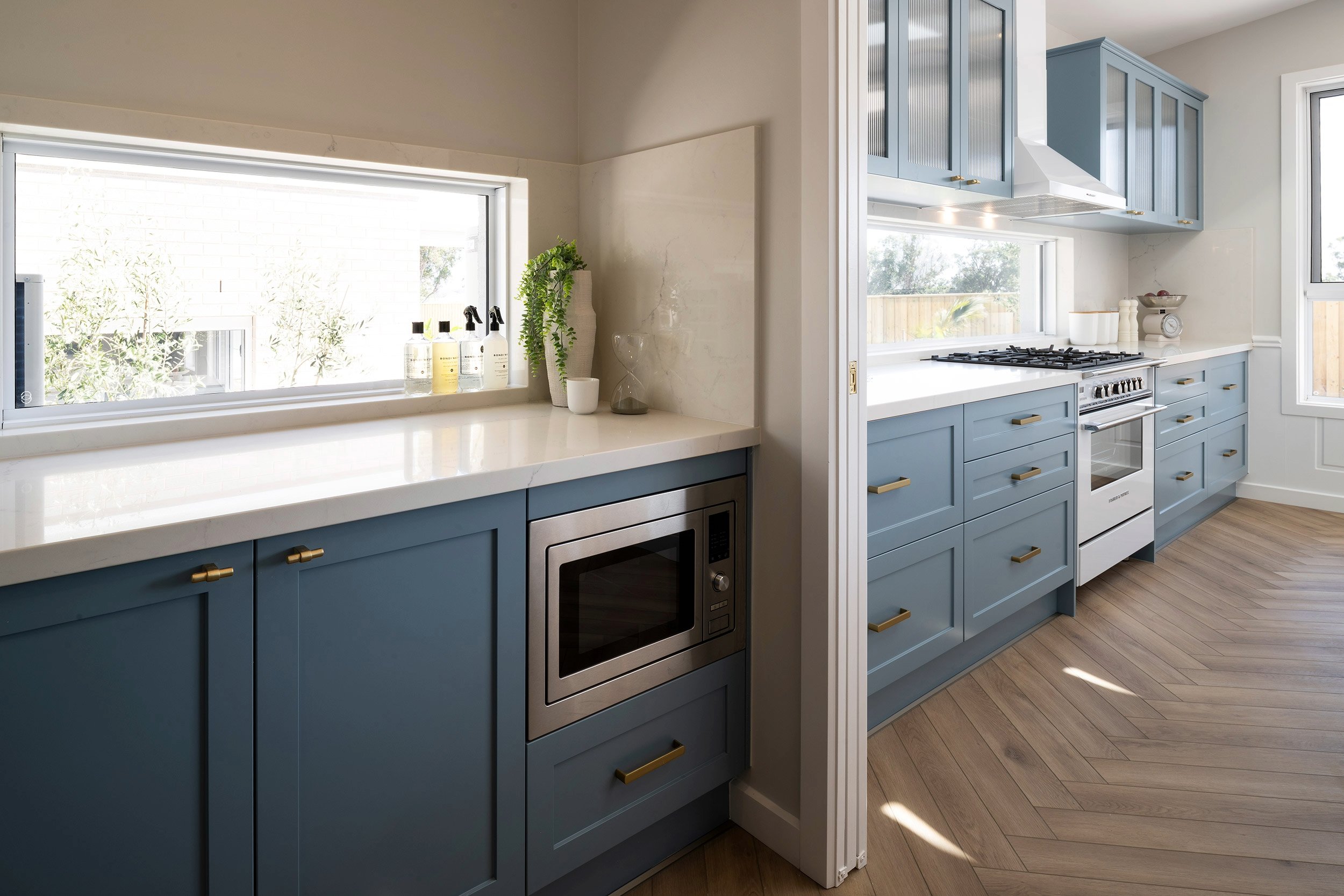 Modern kitchen with blue cabinetry and white countertops, featuring stainless steel appliances, a gas cooktop, built-in microwave, and upper cabinets with glass doors. Large windows with natural light overlook a backyard, and wooden flooring runs throughout the space.
