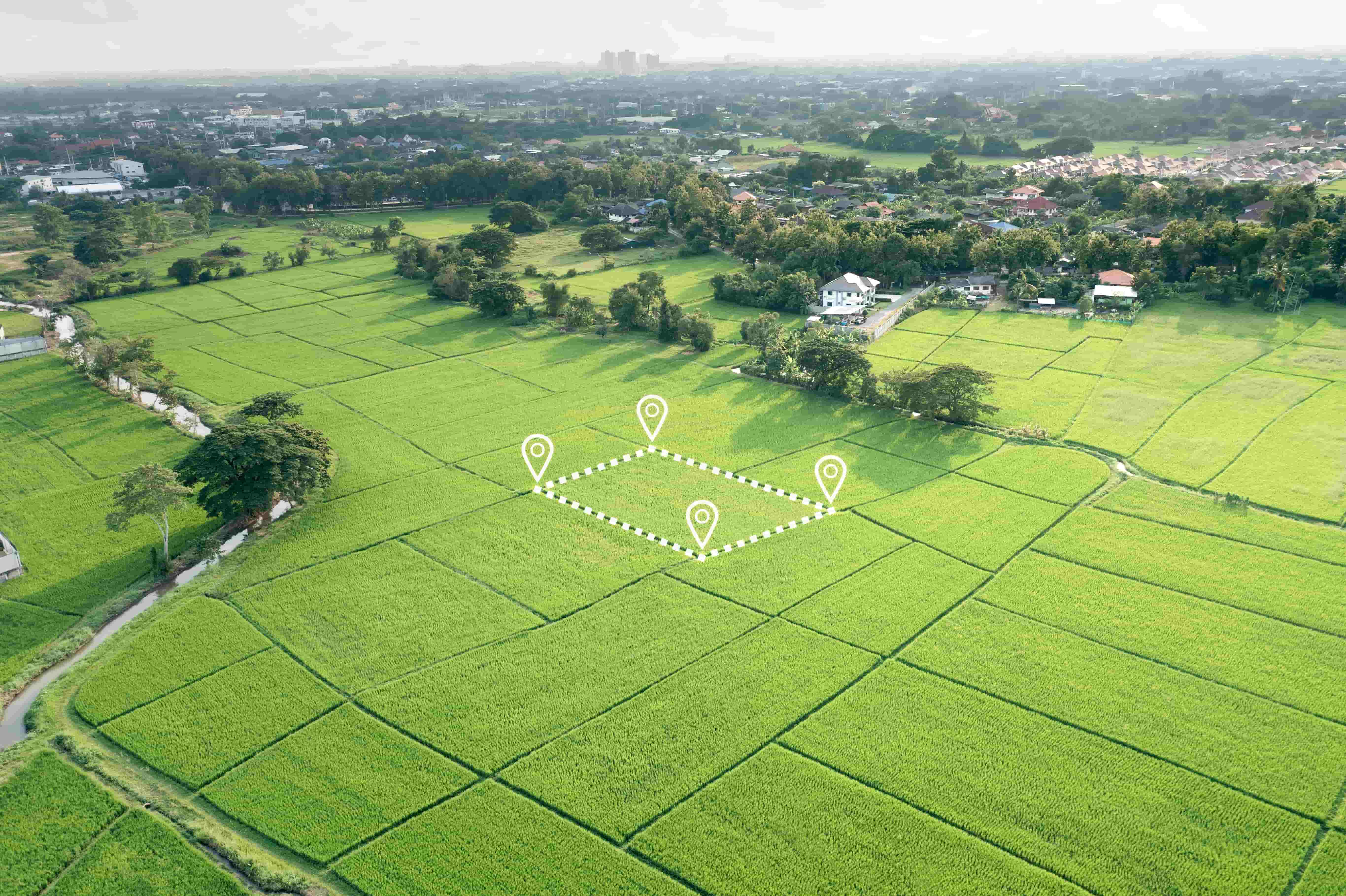 Aerial view of rural farmland with green fields and scattered houses, showing a diamond-shaped outlined plot of land marked with four location pins, representing step 1 of finding a house and land package.