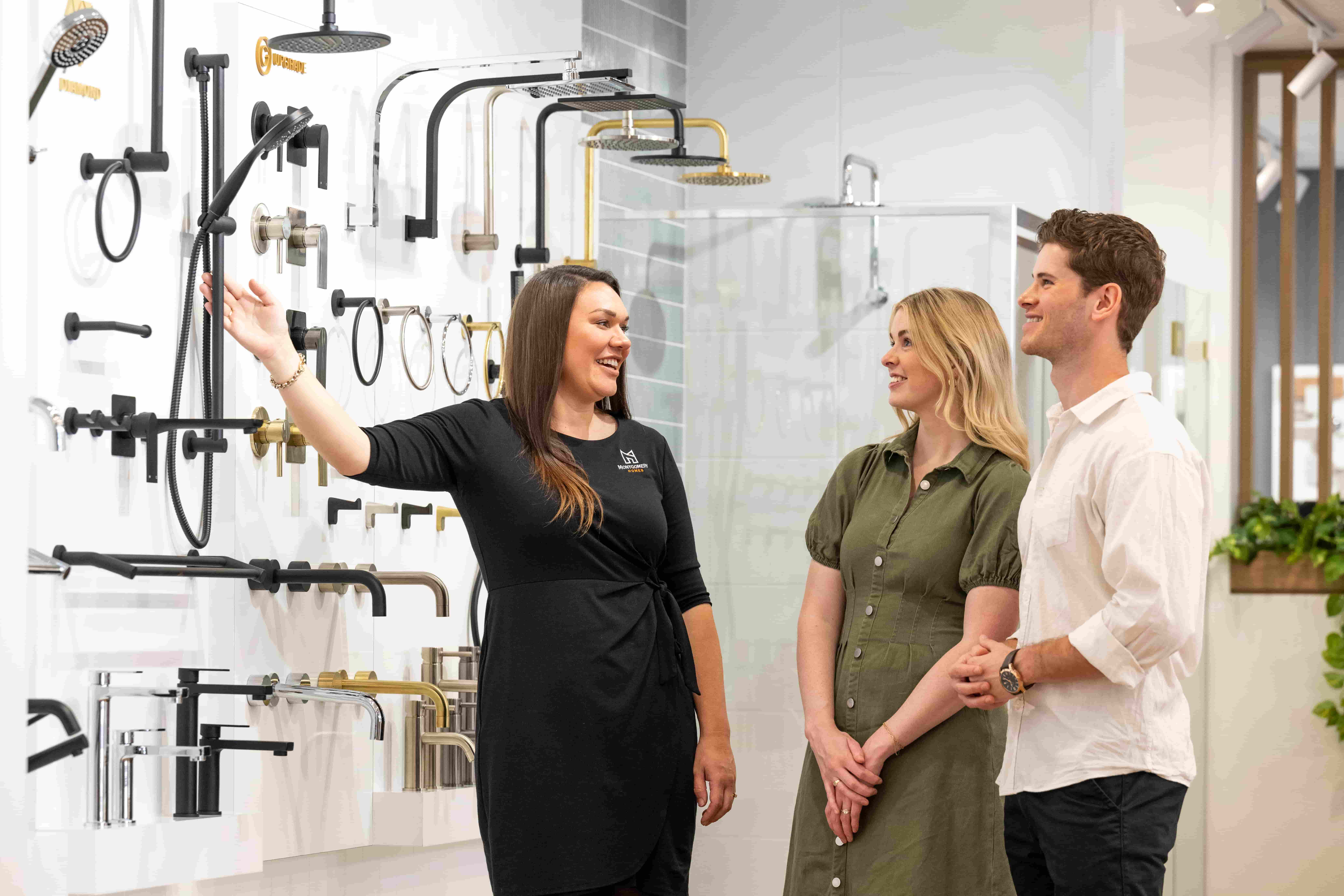 A female sales representative in a black dress gestures toward a bathroom fixtures display wall showing various showerheads and faucets in black and gold finishes, while a couple in casual clothing listens attentively during a showroom appointment.