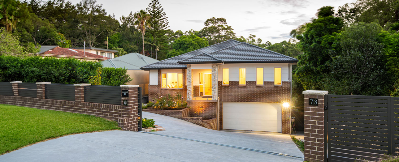 Modern brick and render house at dusk with illuminated windows, dark tile roof, and garage. Property features brick and horizontal slat fencing, concrete driveway, manicured lawn, and mature trees in background. Address number 78 visible on fence pillar.