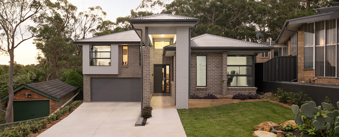 Modern two-story house in Kirrawee with brick and white rendered facade, dark metal roof, gray garage door, and large windows. The front features a concrete driveway, manicured lawn, native plants, and eucalyptus trees. Adjacent properties visible on either side.