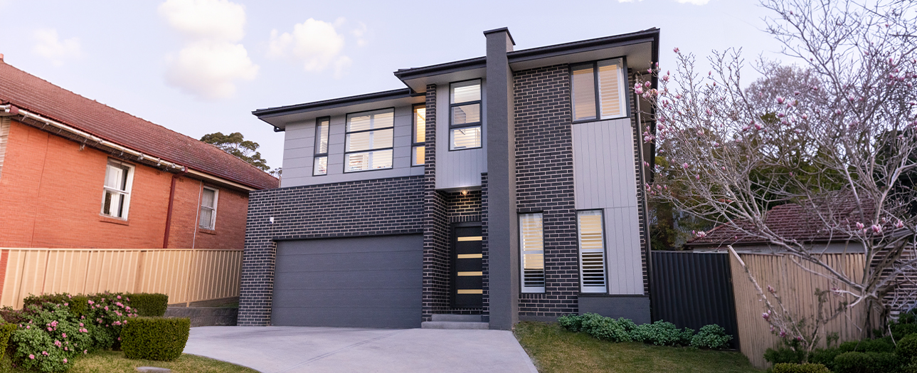 Modern two-story house with dark brick facade and gray metal upper level, featuring a dark garage door, large windows, and contemporary design. Adjacent red brick home visible on left. Bare magnolia tree and landscaping in front yard.