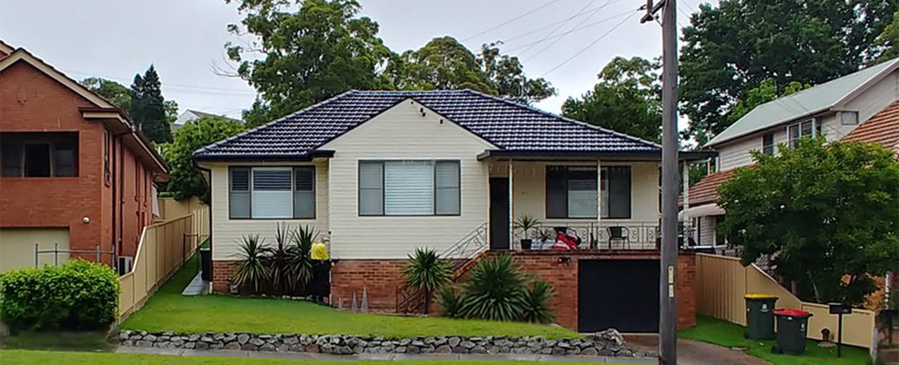 Single-story brick and weatherboard house with dark tiled roof, white horizontal siding upper level, red brick lower level, front porch with metal railing, and landscaped front yard with palm plants and green lawn. Neighboring houses visible on either side.