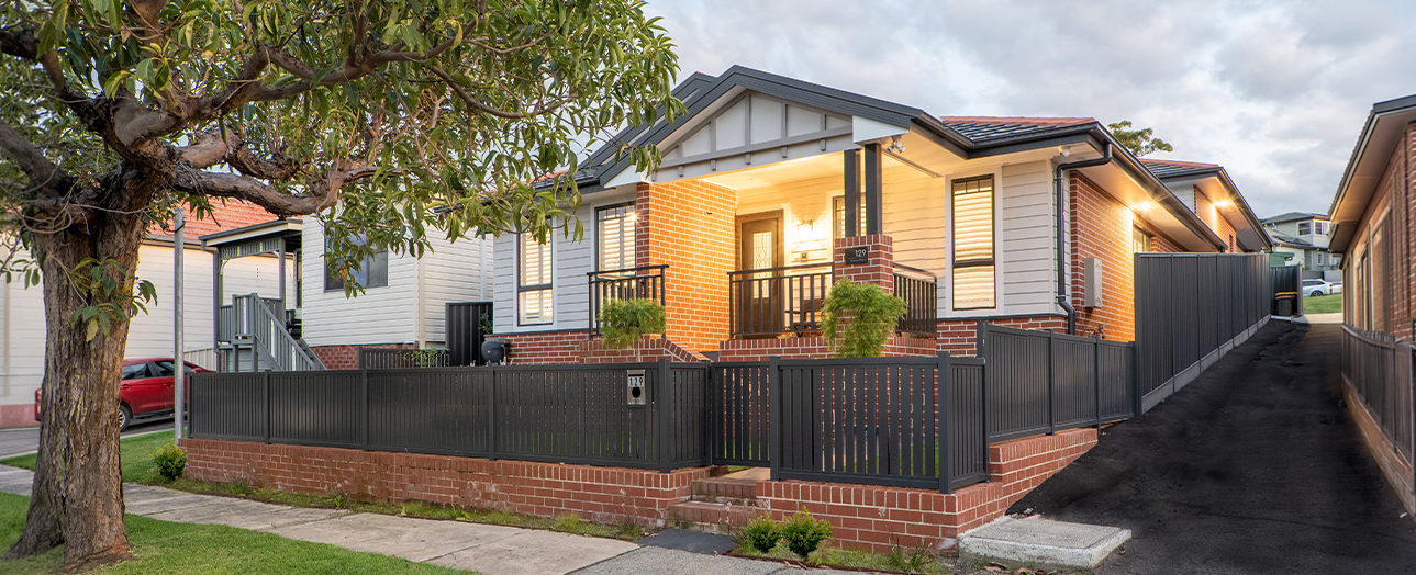 Renovated brick and weatherboard house with modern black metal fencing, illuminated front porch, and manicured garden. Large tree in foreground, driveway to the right, neighboring houses visible, cloudy sky above.