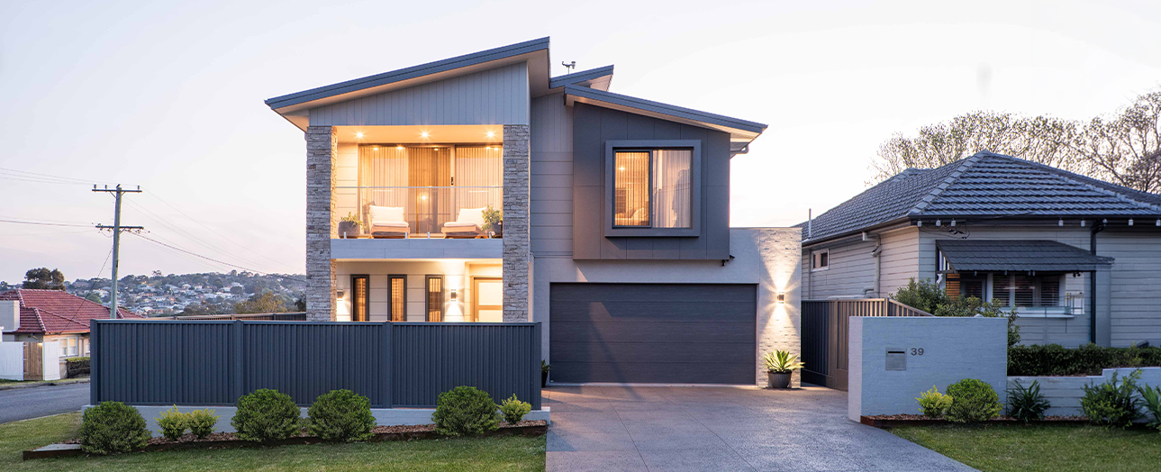 Modern two-story house with dark blue metal cladding and stone accents, illuminated windows, dark garage door, manicured front landscaping, and driveway, positioned between older neighboring houses on a residential street at dusk.