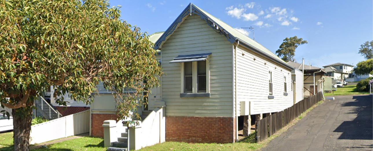 Single-story weatherboard house with cream and light blue exterior, elevated on brick stumps, featuring a peaked roof and front awning. White picket fence and mature tree in overgrown front yard. Residential street with neighboring houses visible in background under clear blue sky.
