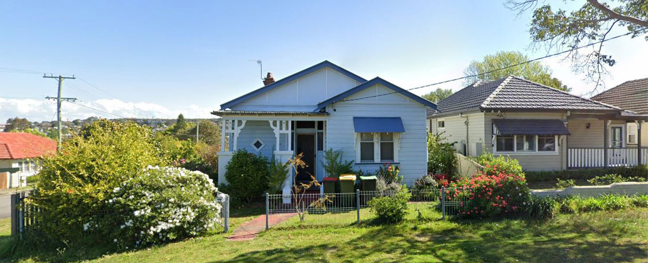 Light blue weatherboard house with dark blue roof and awnings, surrounded by overgrown garden with white flowering shrub on left and red flowers on right, brick front path, metal railings, neighboring houses visible, clear sky, power lines in background.