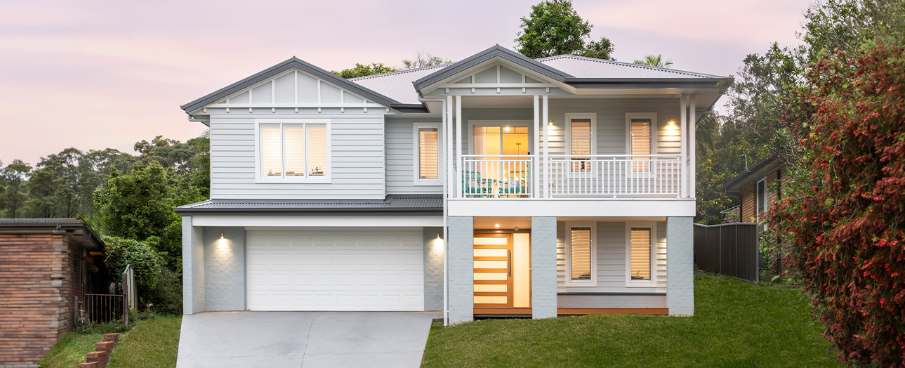 Modern two-story white house with gray roof at dusk, featuring a double garage, front porch with white railings, illuminated windows and entry door, manicured lawn, and surrounding trees and ivy-covered fence.