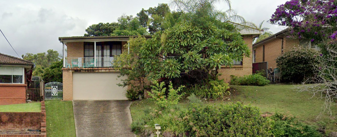 Brick residential house with overgrown vegetation covering the front facade and landscaping. The two-story home features a garage, upper balcony, and is surrounded by dense green plants and trees. Adjacent brick houses visible on either side. Overcast sky above. The property appears unkempt with wild growth obscuring the home's features.