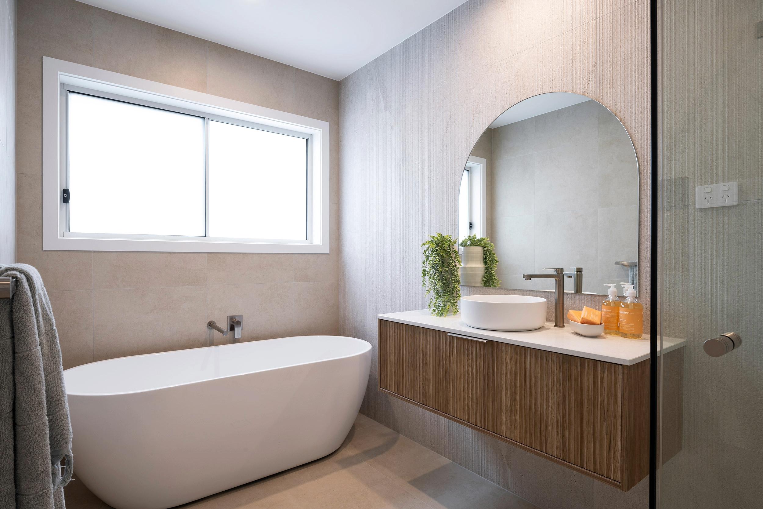 Luxe resort bathroom featuring a freestanding white oval bathtub on the left, a large arched mirror above a wooden vanity with white vessel sink on the right, neutral beige and cream tiled walls, a horizontal window with white frame, towel rack, potted plant, bath amenities, and glass shower enclosure.