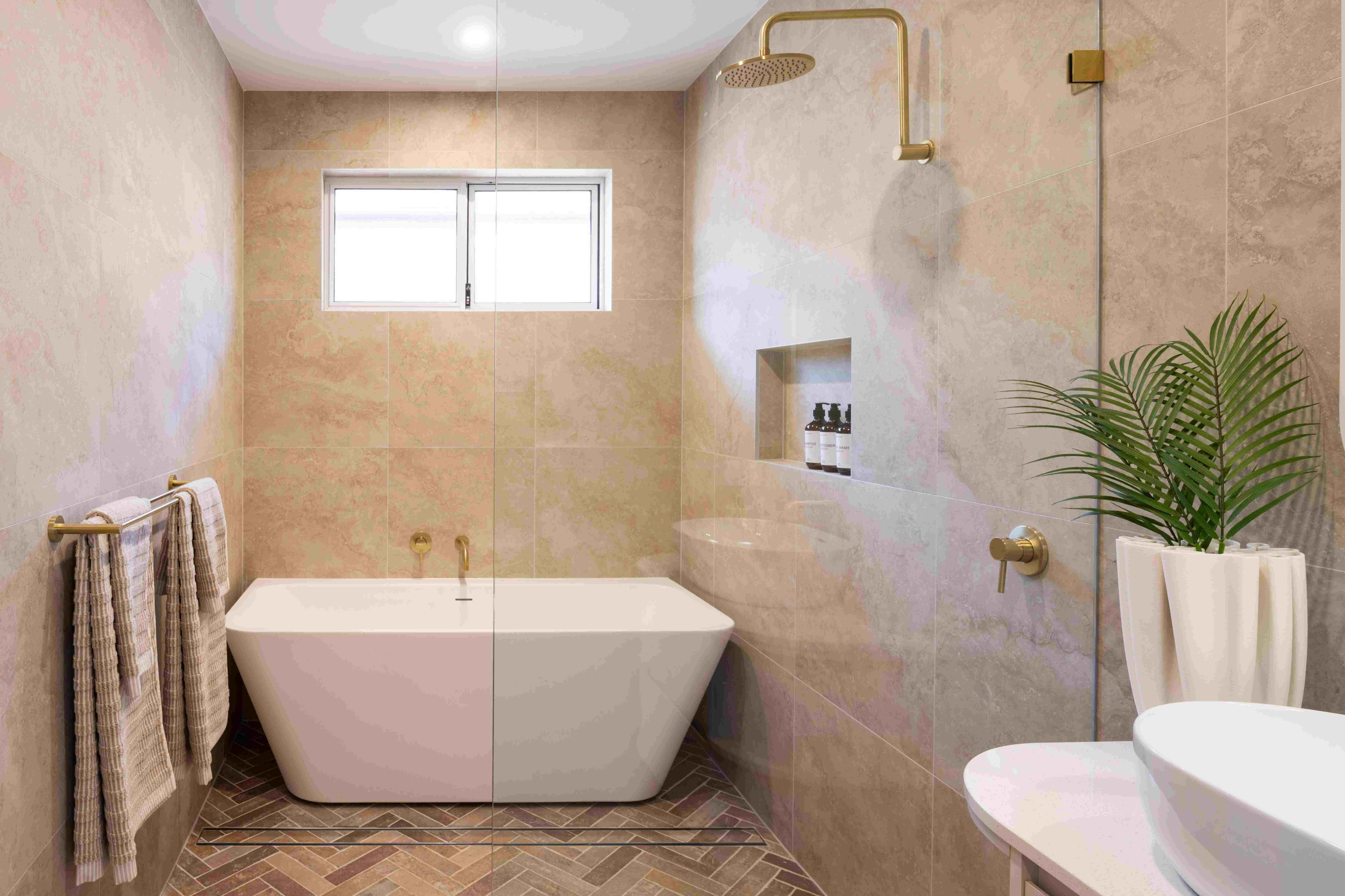 Luxurious bathroom with a freestanding white soaking tub centered in the room, surrounded by warm beige marble tiles. Brass rainfall showerhead and fixtures are mounted above the tub. A frameless glass shower enclosure is visible on the right side with built-in niche storage. A small window provides natural light above the tub. Herringbone wood-pattern flooring, gold towel racks with striped towels, a potted green