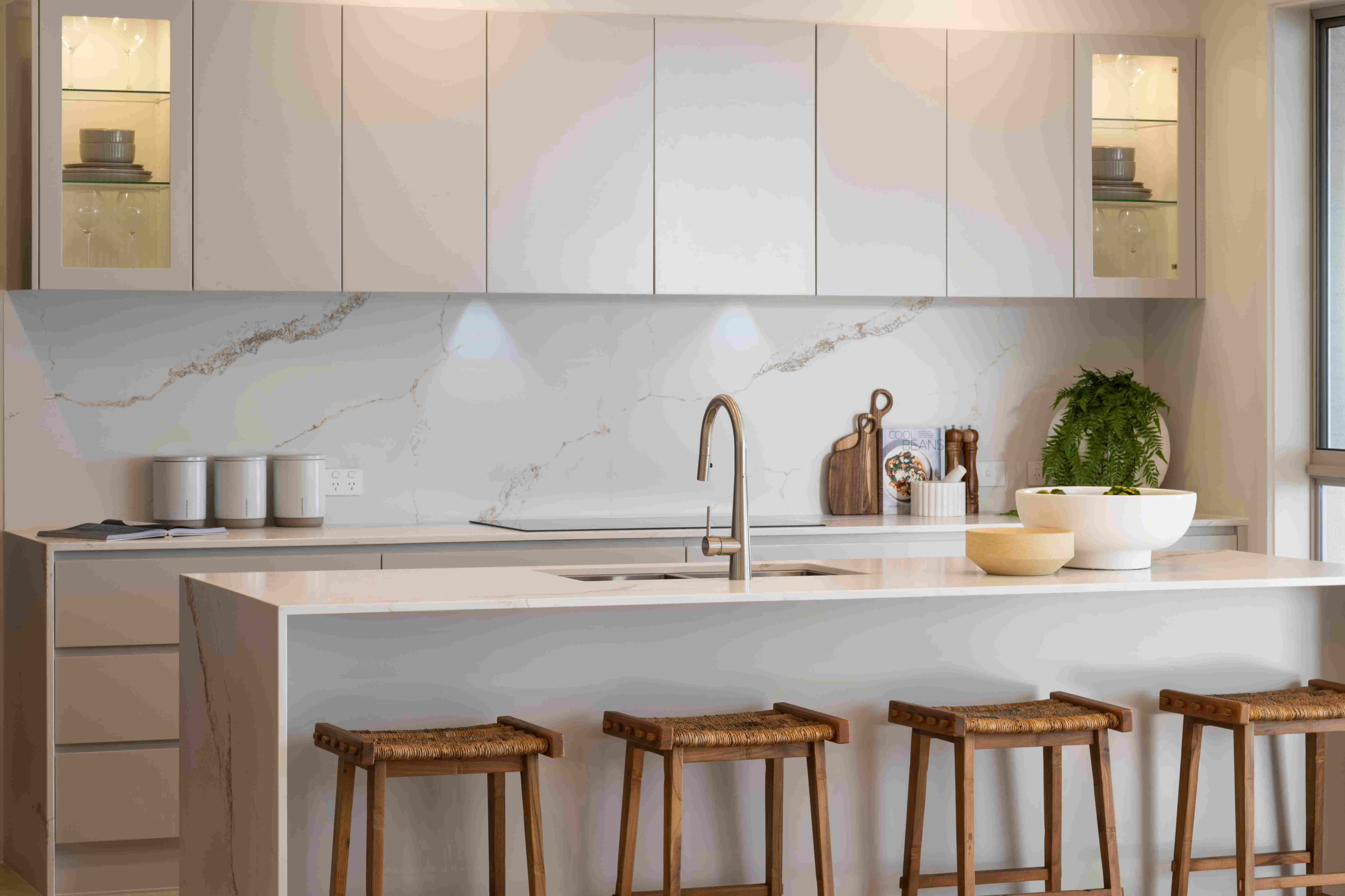 Luxe resort kitchen with white cabinetry, marble backsplash with gold veining, and waterfall island countertop. Bronze faucet, wooden woven bar stools, and decorative accessories including potted ferns and stainless steel containers. Illuminated upper cabinets with display shelving visible on sides.