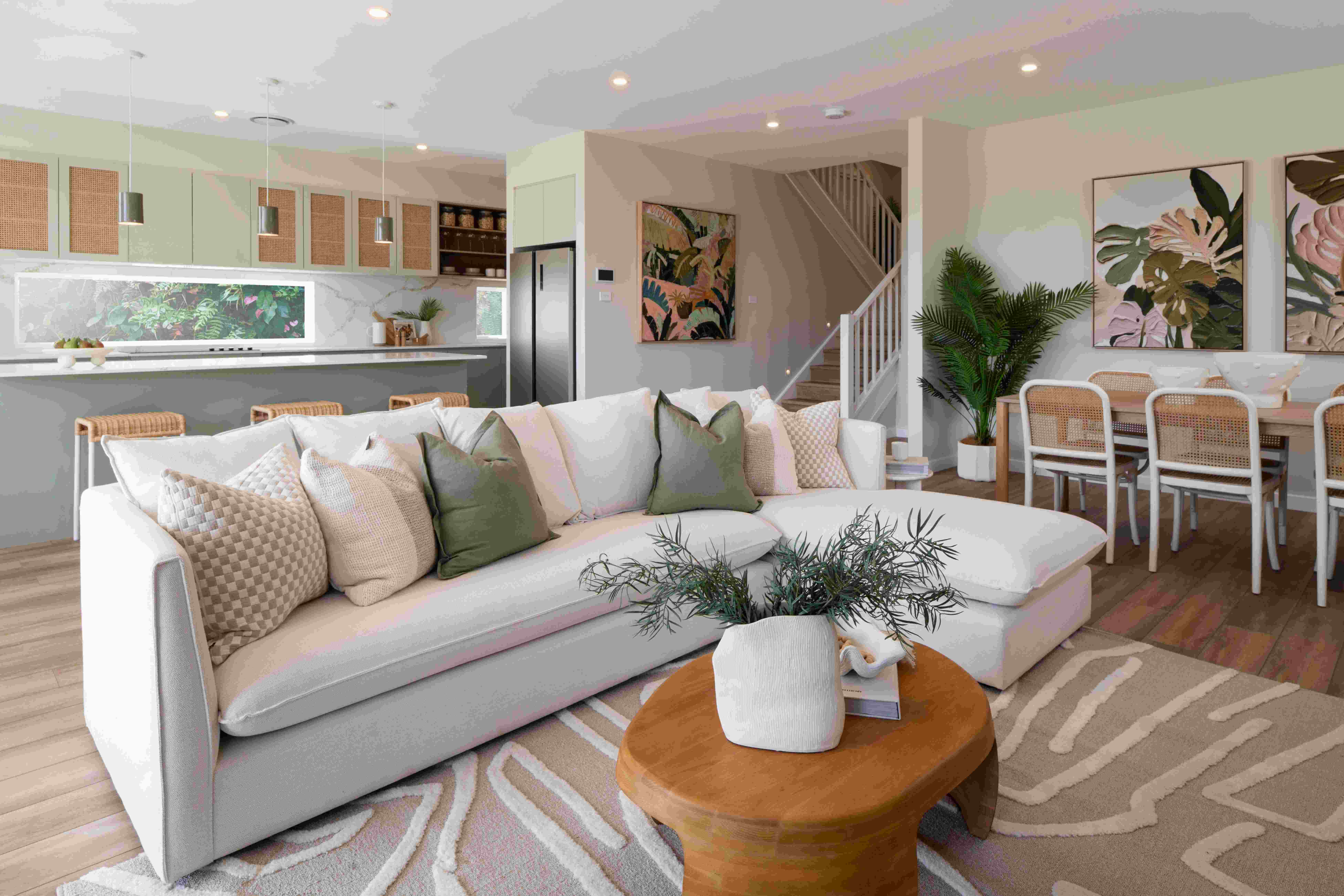 Luxe resort living room with light green and cream walls, featuring a large white sectional sofa with patterned pillows, wooden round coffee table with potted plant, open-concept kitchen with mint cabinets and stainless steel appliances, dining area with rattan chairs, botanical wall art, indoor plants, and staircase leading to upper level.