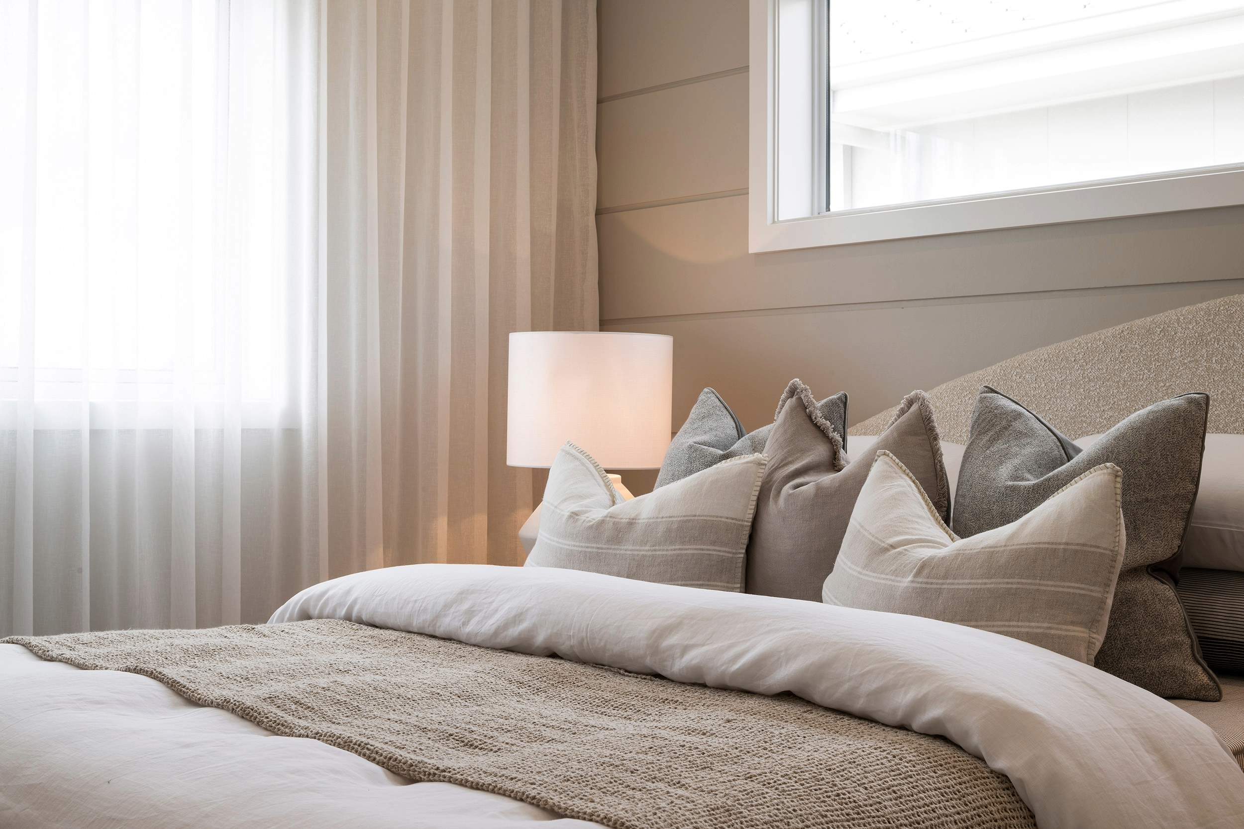 Luxe resort bedroom with neutral tones featuring a bed with white pillows and decorative gray and beige cushions, a textured throw blanket, a soft-glowing bedside lamp, floor-to-ceiling windows with layered curtains, and minimal modern décor in warm, calming tones.