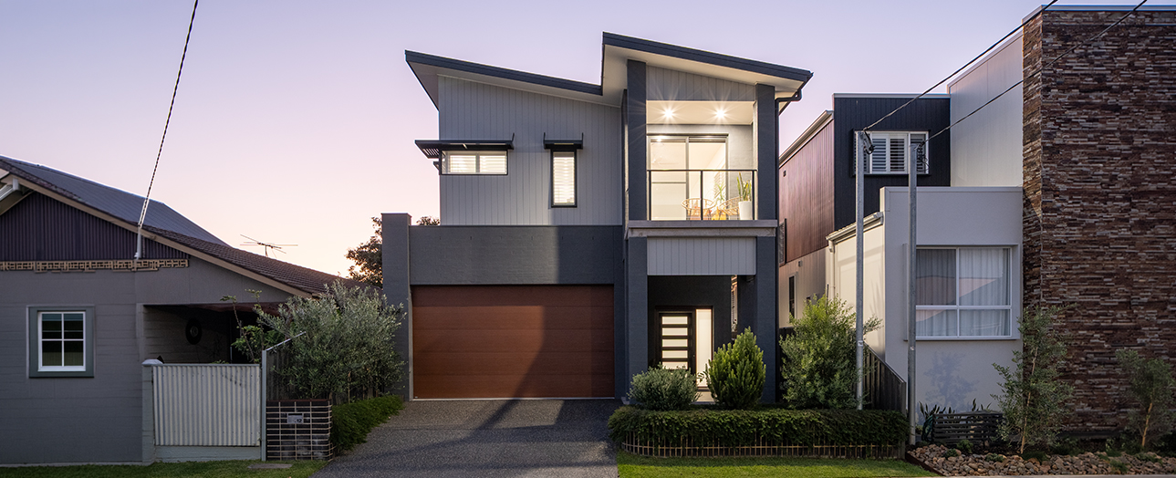 Modern two-story residential home with dark gray and white exterior, featuring a copper-brown garage door, large illuminated upper windows, and contemporary architectural design. Flanked by older neighboring houses on a tree-lined street at dusk.