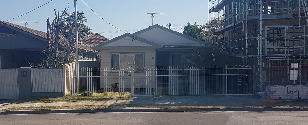 Single-story residential house with gray exterior and peaked roof, enclosed by white metal security fencing across the front. Left side shows white corrugated metal fence, right side shows multi-story building under construction with metal scaffolding. Utility poles and power lines visible overhead. Clear blue sky, sparse vegetation in yard.