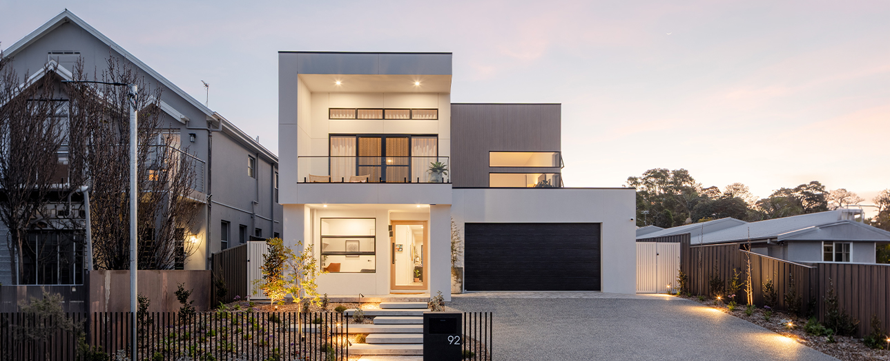 Modern two-story white house with dark accents at dusk, featuring illuminated windows, a black garage door, concrete driveway, and metal fencing. An older gray house is visible on the left side of the image.