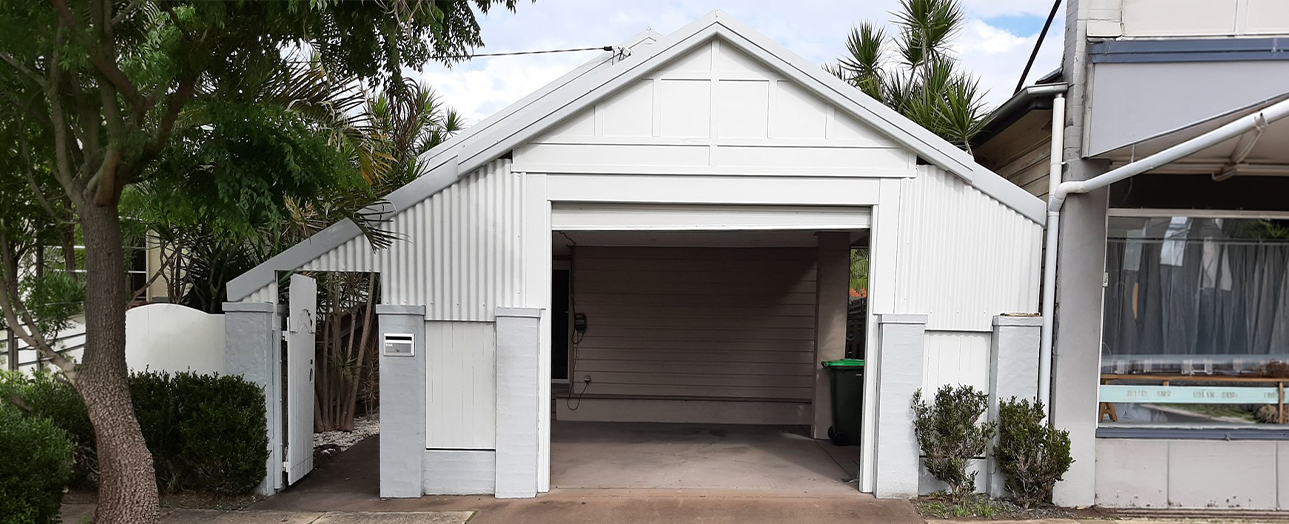 White corrugated metal garage with open brown garage door, surrounded by palm trees and tropical vegetation, with neighboring houses visible on either side and a driveway in front.