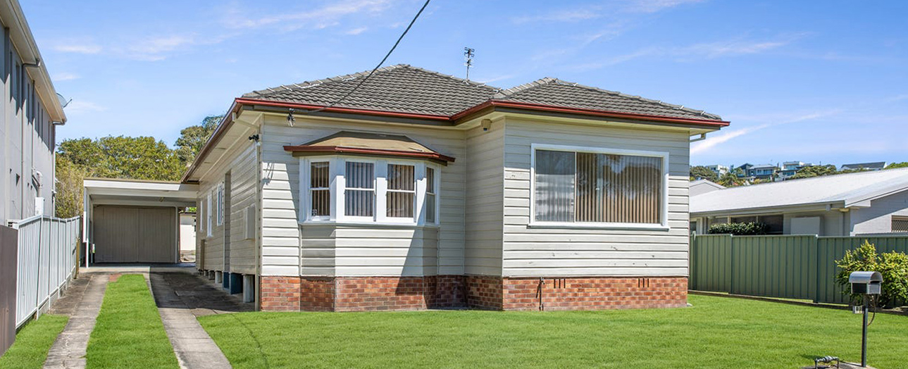 Single-story brick and weatherboard house with dual gabled roof, set on stilts with brick foundation. Features front windows with vertical blinds, attached garage on left side, manicured lawn, and neighboring houses visible. Clear blue sky with light clouds overhead.