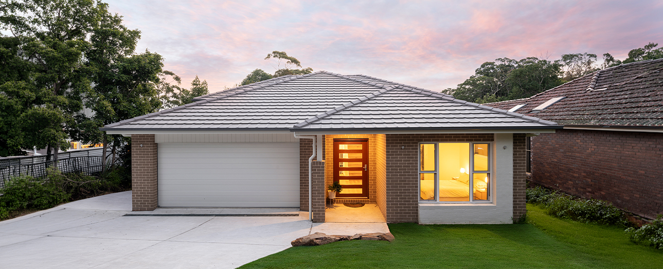 Modern single-story brick house at dusk with illuminated entry door and windows, featuring a new gray metal roof, white garage door, concrete driveway, and manicured lawn surrounded by trees.